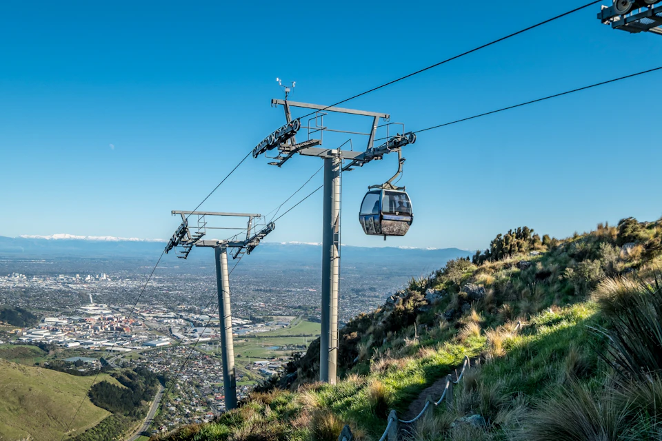 A gondola lift ascending a hillside with green vegetation, overlooking a cityscape under a clear blue sky.