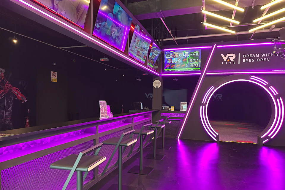 A modern VR gaming lounge with neon purple lighting, bar stools, screens displaying games, and a VR Live logo on the wall.