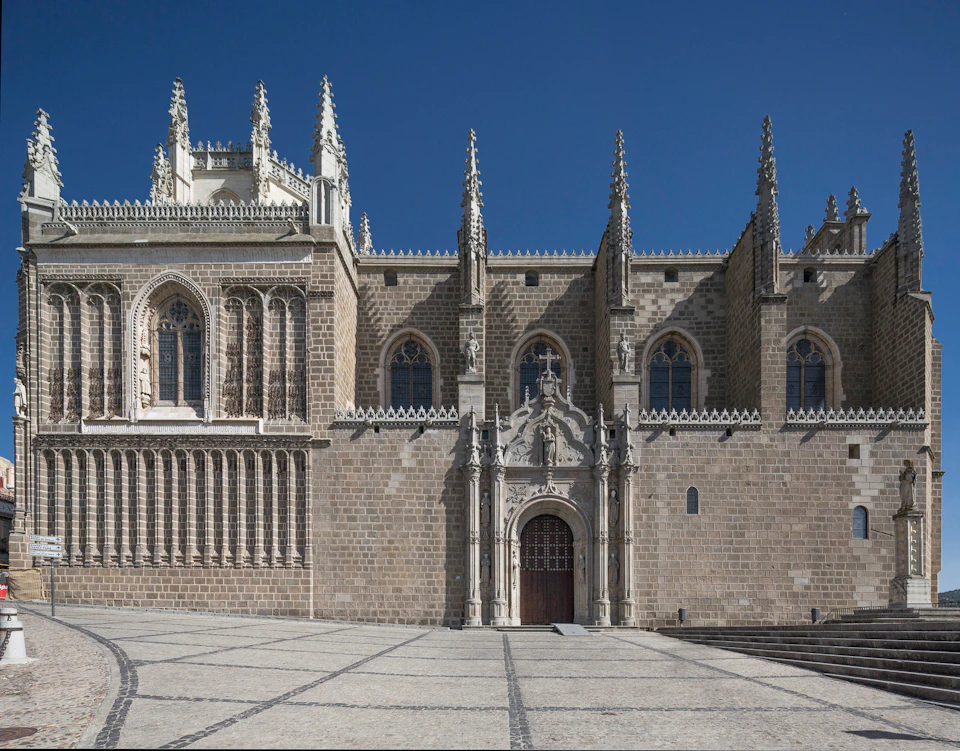 Gothic-style stone building with ornate detailing, arched windows, and spires under a clear blue sky. Courtyard in the foreground.