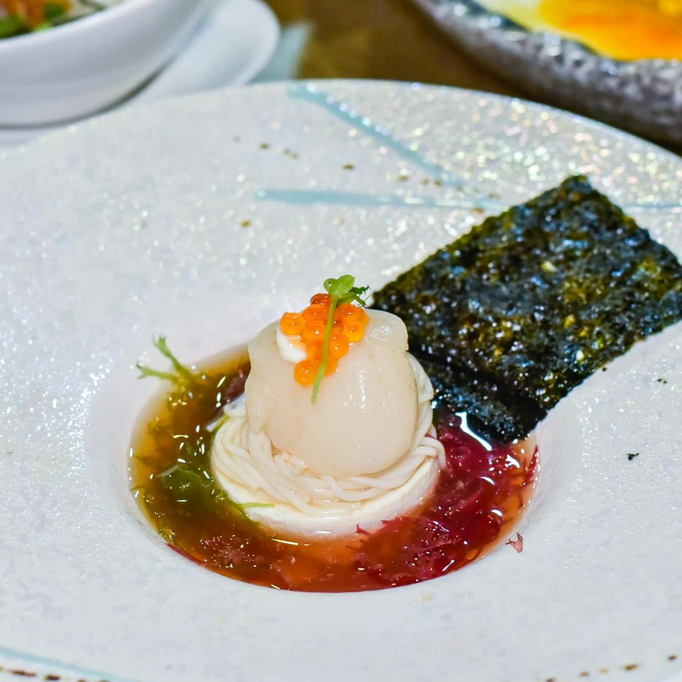 A plated dish featuring a scallop topped with orange roe, seaweed, and colorful sauces presented on a white plate.