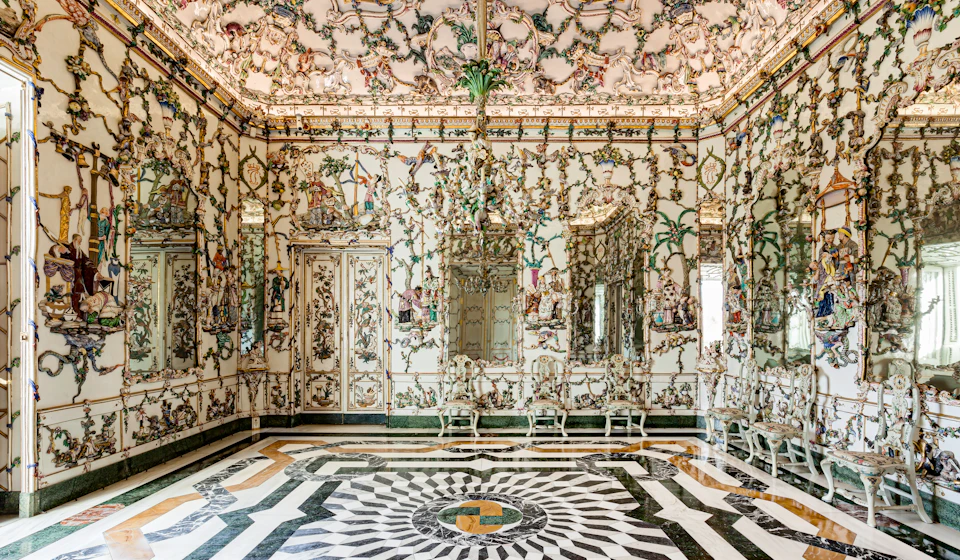 Ornate room with intricate wall and ceiling designs, mirrors, chandeliers, white chairs, and a black and white patterned floor.