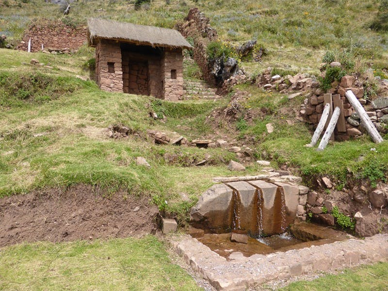 Small stone hut with a thatched roof, situated on a grassy hillside. Water flows from a stone fountain in front of the hut.