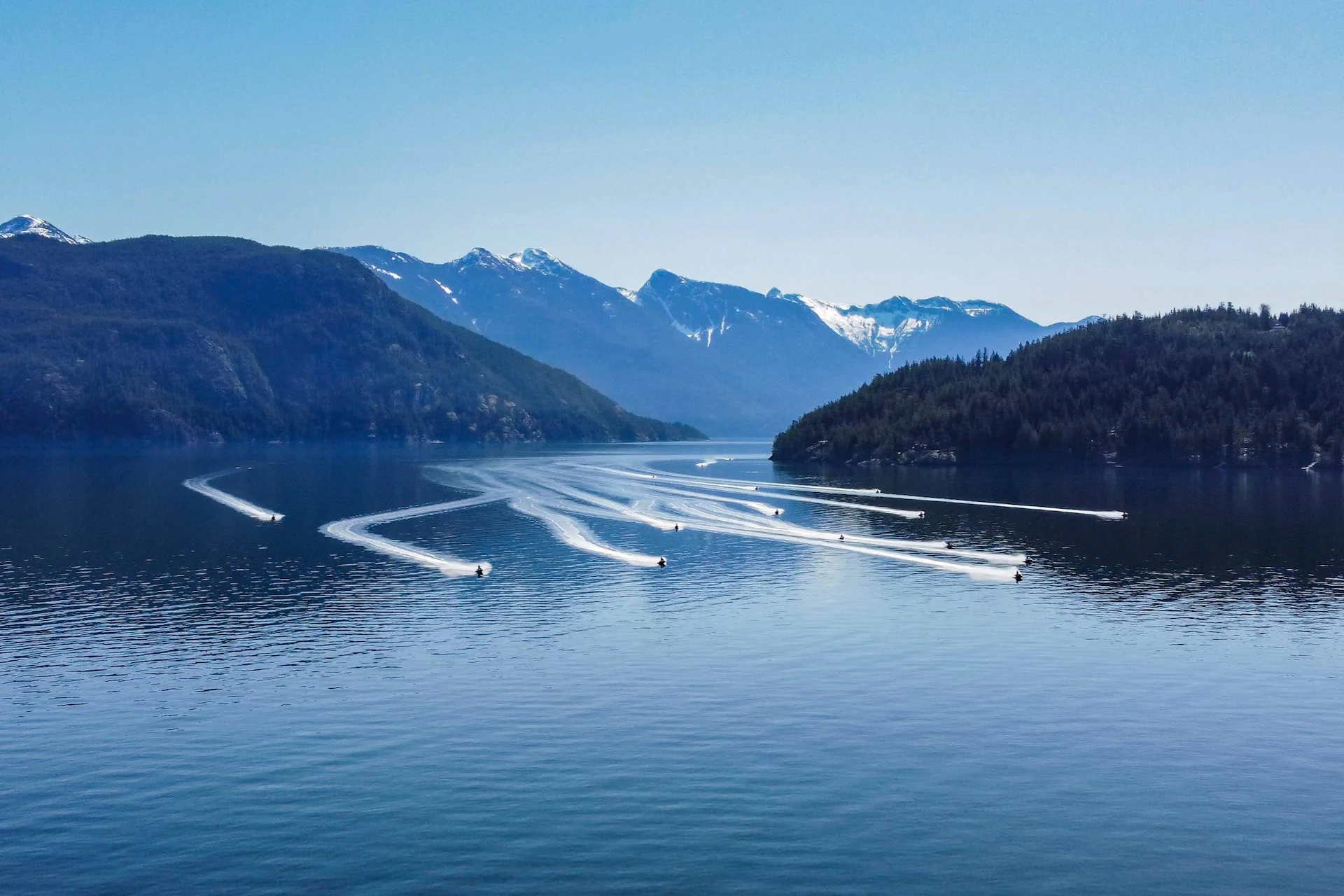 Multiple boats create white trails on a calm lake surrounded by mountains with snowy peaks under a clear blue sky.