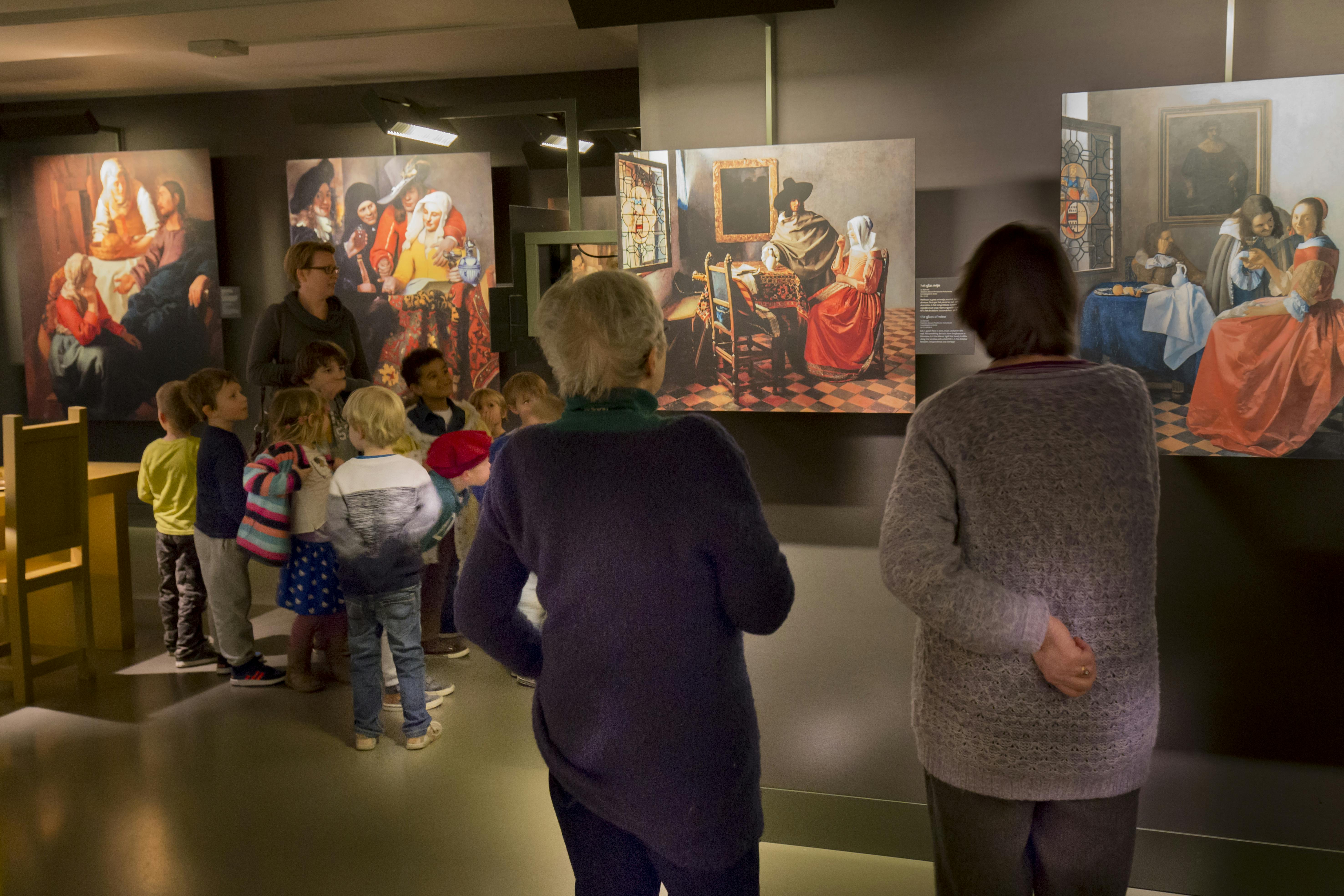 Crianças e adultos observam pinturas num museu, com obras de arte pormenorizadas que retratam cenas históricas nas paredes.