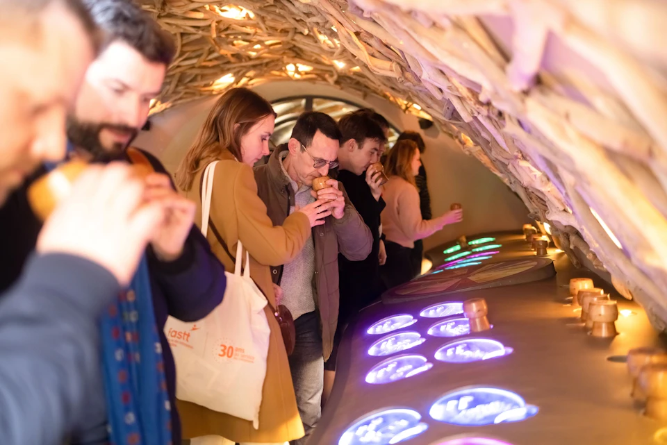 People standing in a line under a wooden arch, holding objects to their noses, possibly engaging in a sensory activity.