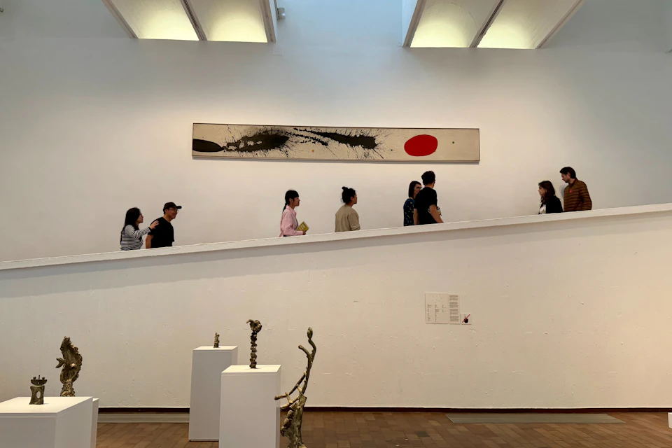 People walking along a ramp in an art gallery with sculptures and a large abstract painting on the wall.