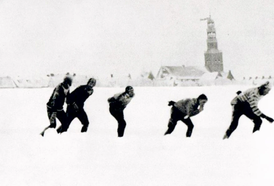 Four people ice skating in a snowy landscape with buildings and a tower in the background.