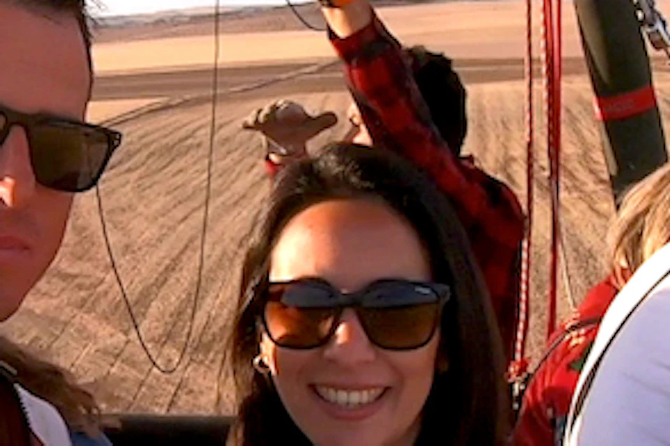 Two people in sunglasses smiling inside a hot air balloon basket, floating above a vast desert landscape.