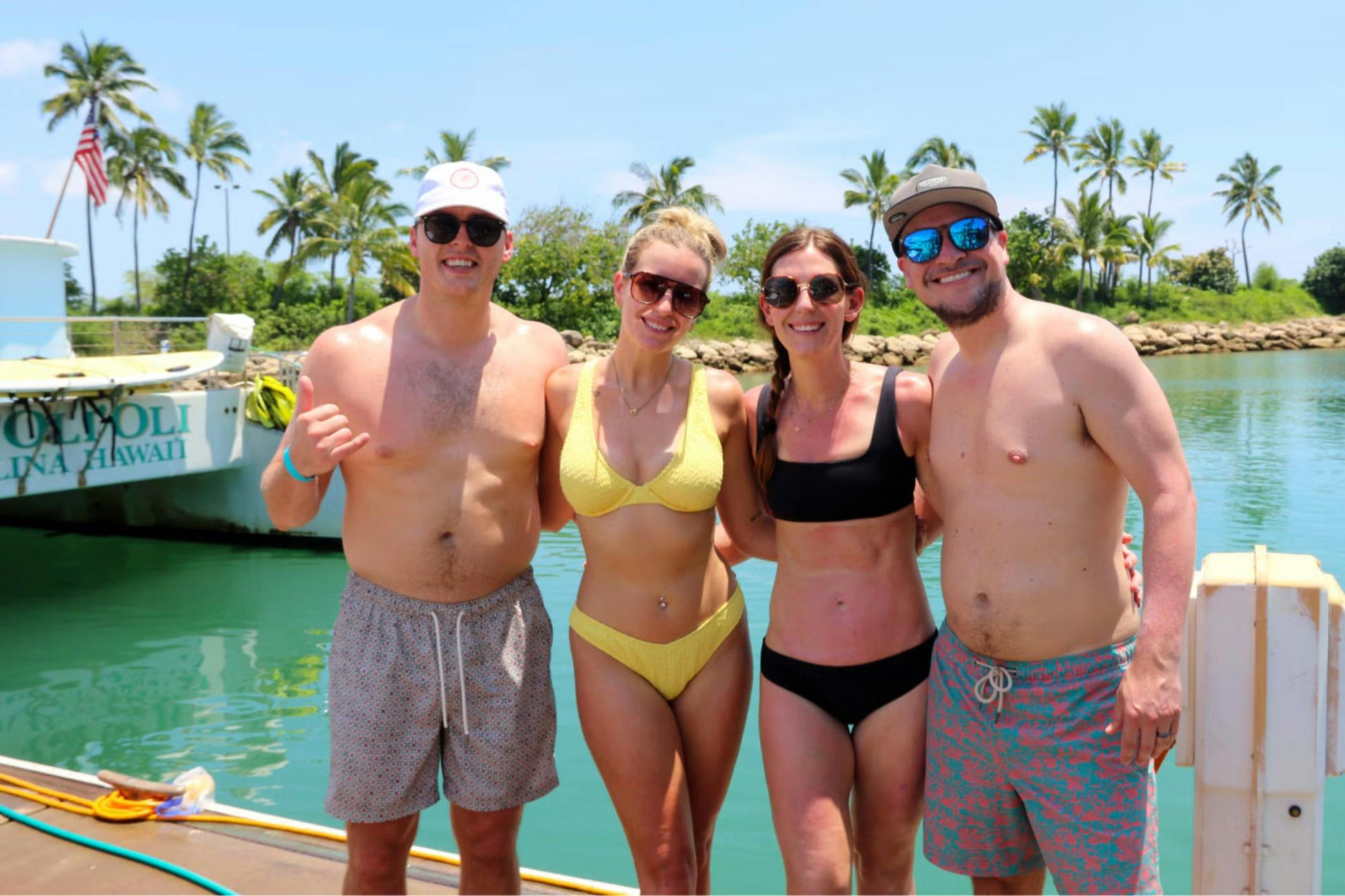 Four people in swimsuits smiling and posing near a body of water with palm trees and a clear sky in the background.