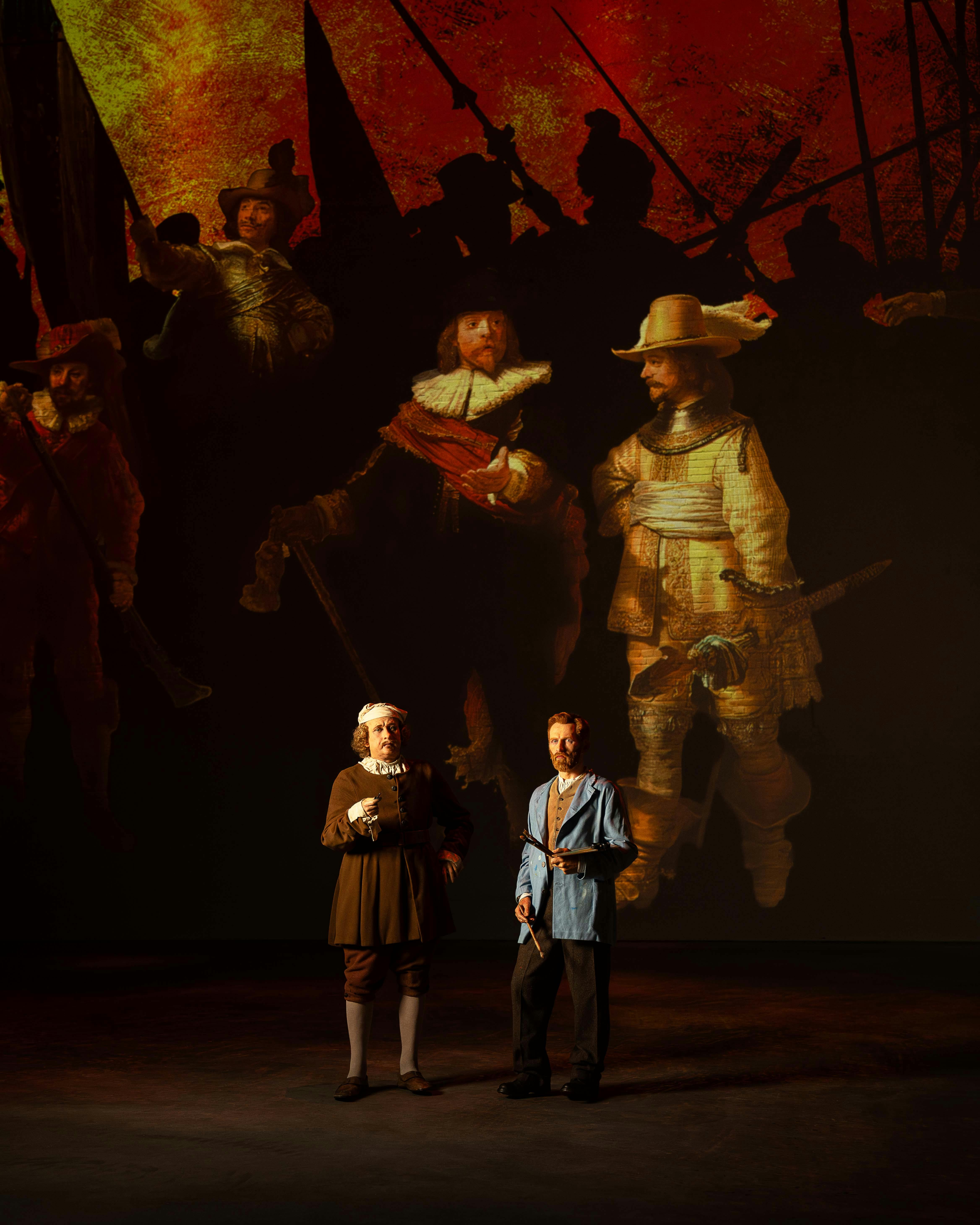 Two men in historical costumes stand in front of a large projection of classical art featuring soldiers.