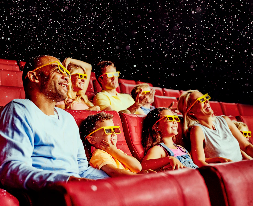 Group of people wearing 3D glasses, sitting in red theater seats, smiling and looking up at floating particles in the air.