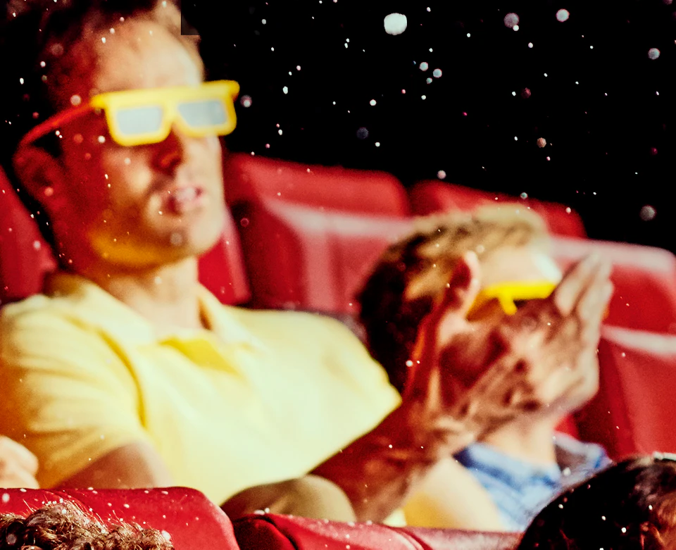 Group of people wearing 3D glasses, sitting in red theater seats, smiling and looking up at floating particles in the air.