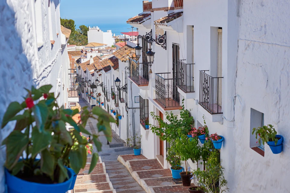 A picturesque narrow street lined with white buildings, wrought-iron balconies and potted plants, offering a view of the sea.