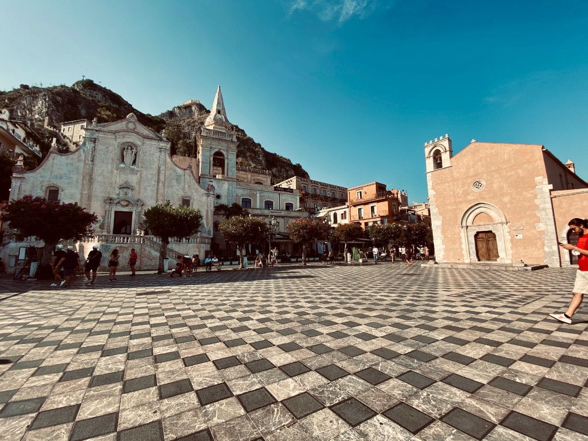 一个城镇广场，有华丽的教堂、其他历史建筑、树木，人们在蓝天下行走。