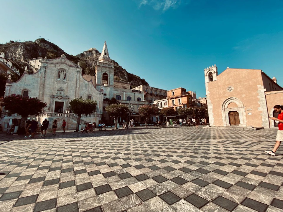 A town square with an ornate church, other historic buildings, trees, and people walking under a blue sky.