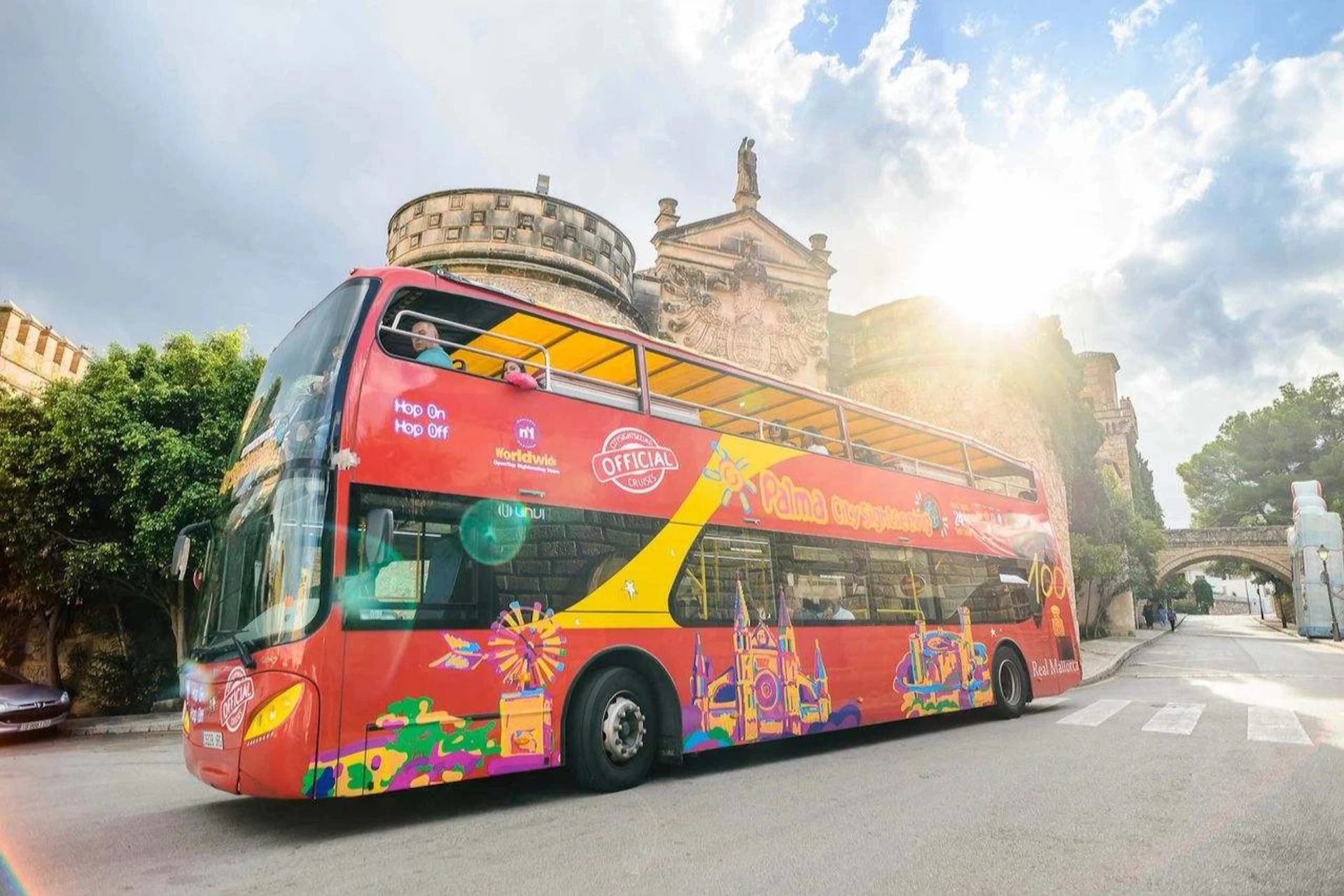 A red double-decker tour bus with colorful graphics on the side, parked near historic buildings under a partly cloudy sky.