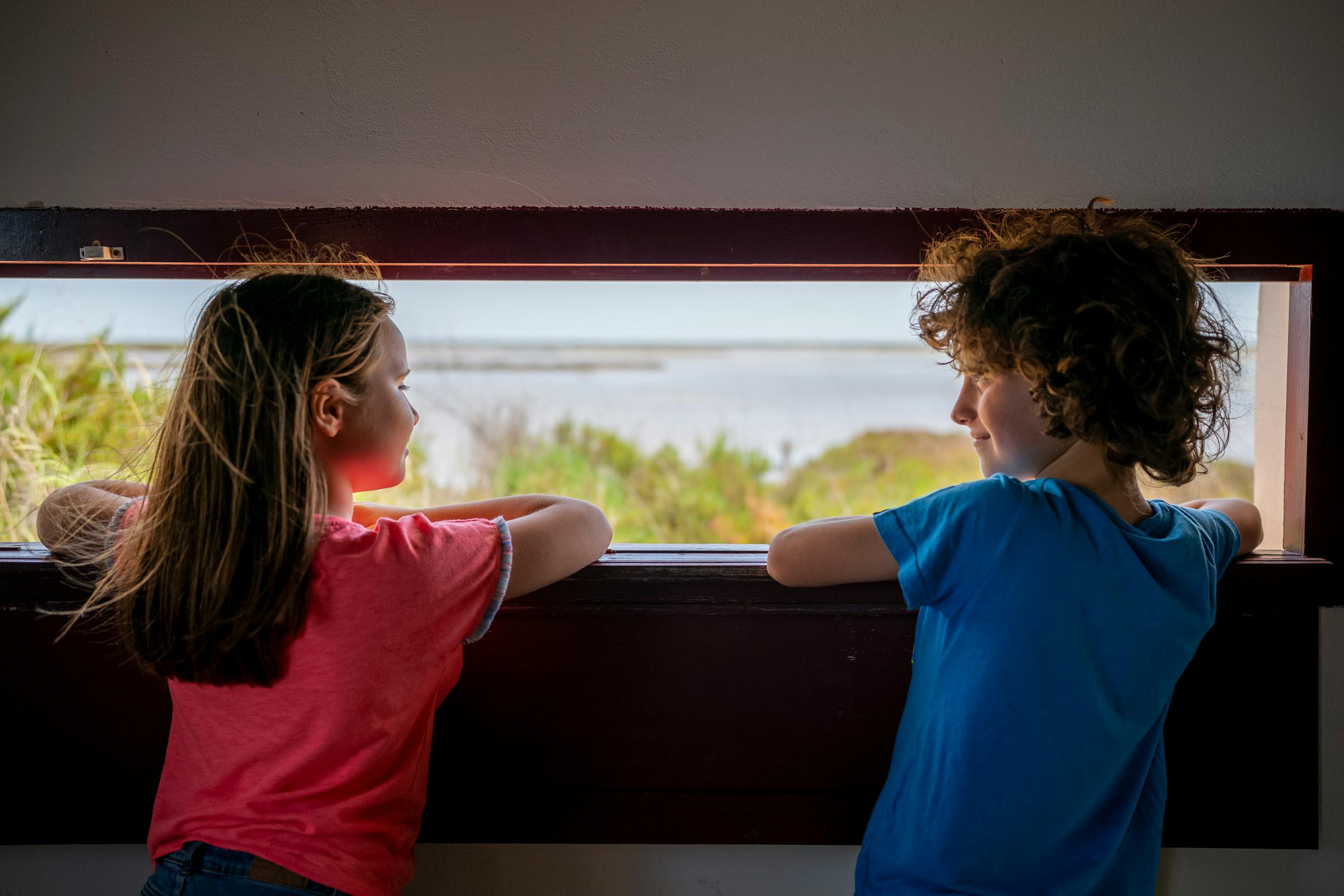 Δύο παιδιά κοιτάζουν έξω από ένα ξύλινο παράθυρο ένα τοπίο με νερό και πράσινο. Το ένα παιδί φορά μπλε πουκάμισο και το άλλο κόκκινο.