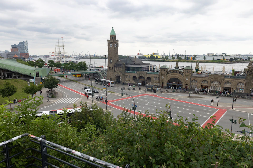 A clock tower and brick building next to a busy intersection overlook a harbor with cranes and ships under a cloudy sky.