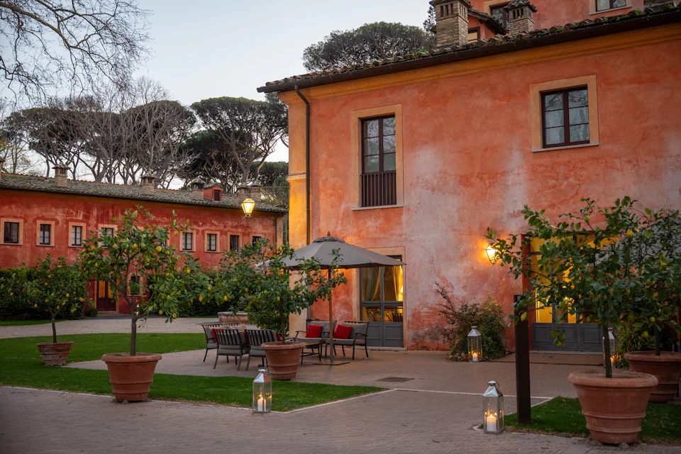 A courtyard with a building featuring orange walls, outdoor seating, potted plants, lanterns, and trees in the background.