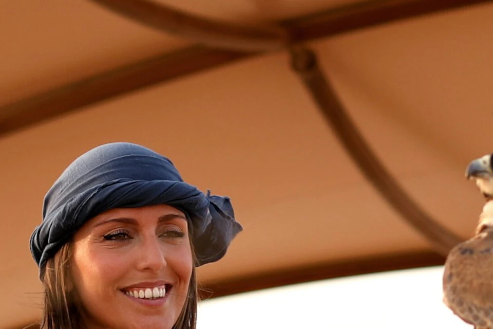 Three people smile under a canopy. One man holds a falcon wearing a hood on a gloved hand. Desert landscape in the background.