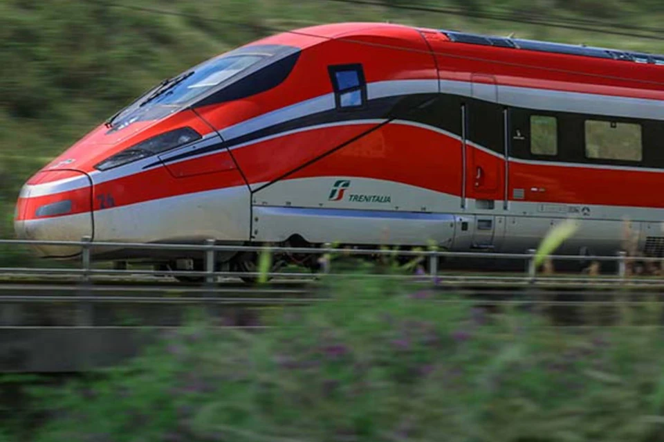 A red and white high-speed train labeled Trenitalia traveling on a railway through a green landscape.