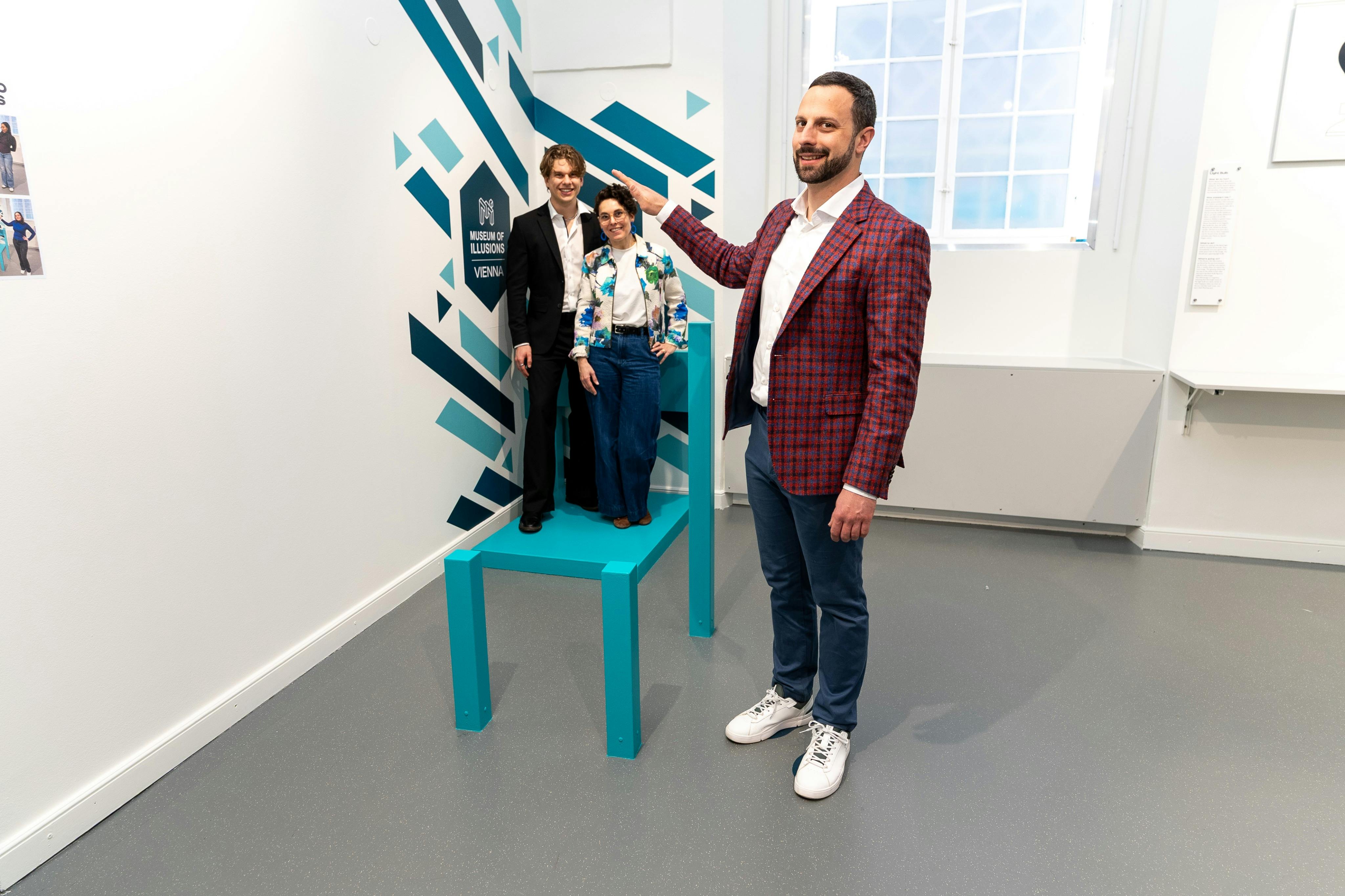 En person i en rød jakke står ved siden af en stor blågrøn stol, med to personer siddende på stolen i en museumsudstilling.