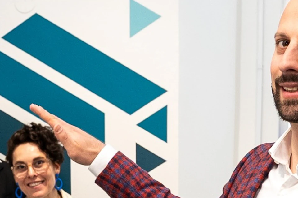 A person in a red jacket stands beside a large teal chair, with two people sitting on the chair in a museum exhibit.