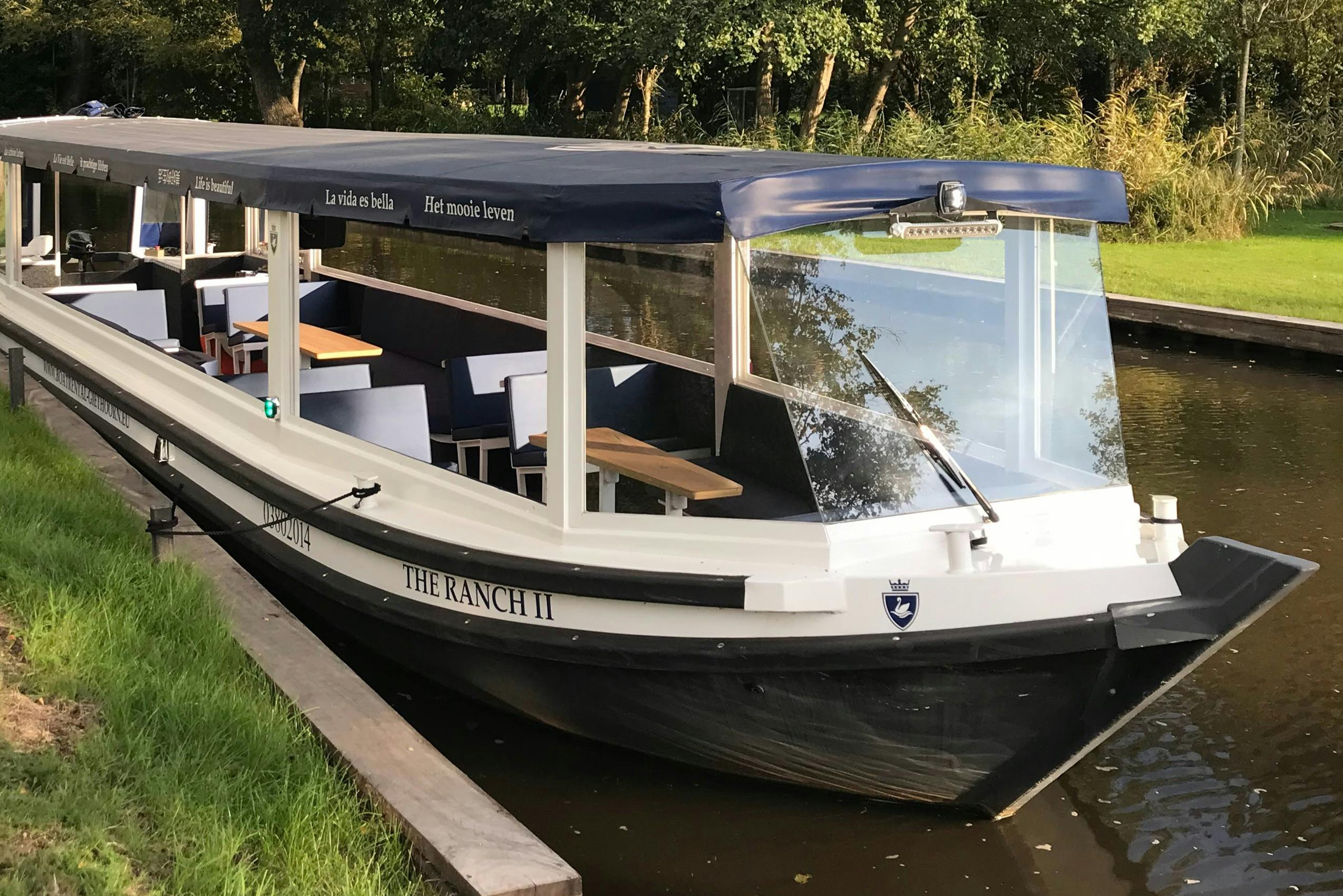 A covered canal boat named "The Ranch II" docked beside a grassy bank, surrounded by calm waters and lush trees.