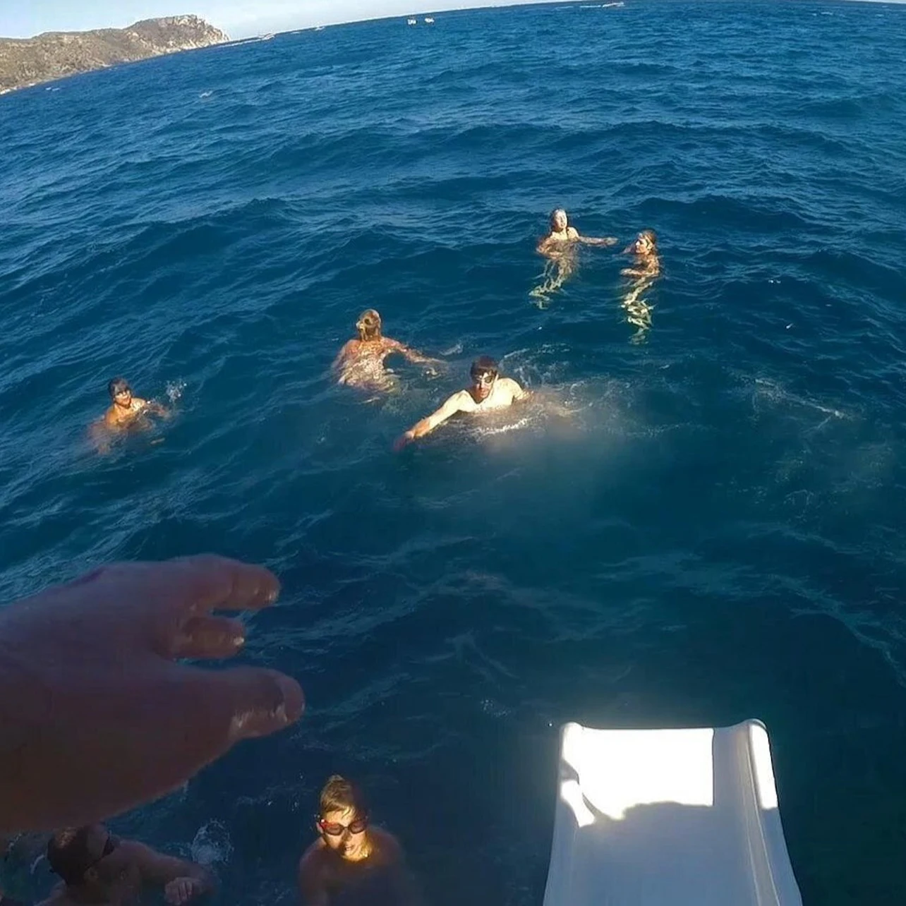 Several people swimming in blue ocean water near a boat with a white slide, with distant land visible on the horizon.