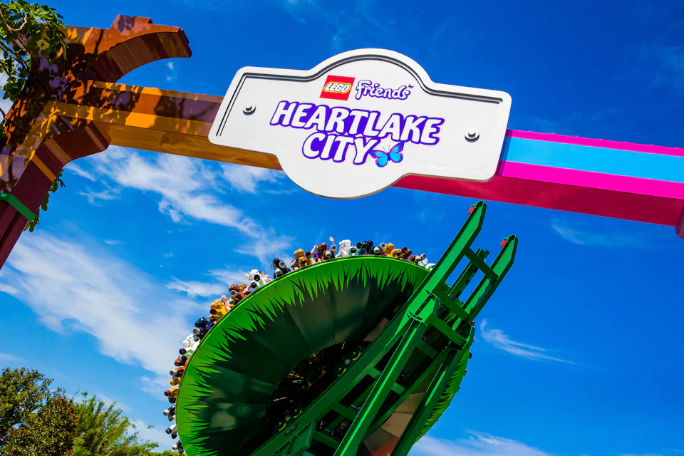 Entrance sign for "LEGO Friends Heartlake City" with a rollercoaster loop and riders in the background, set against a blue sky.