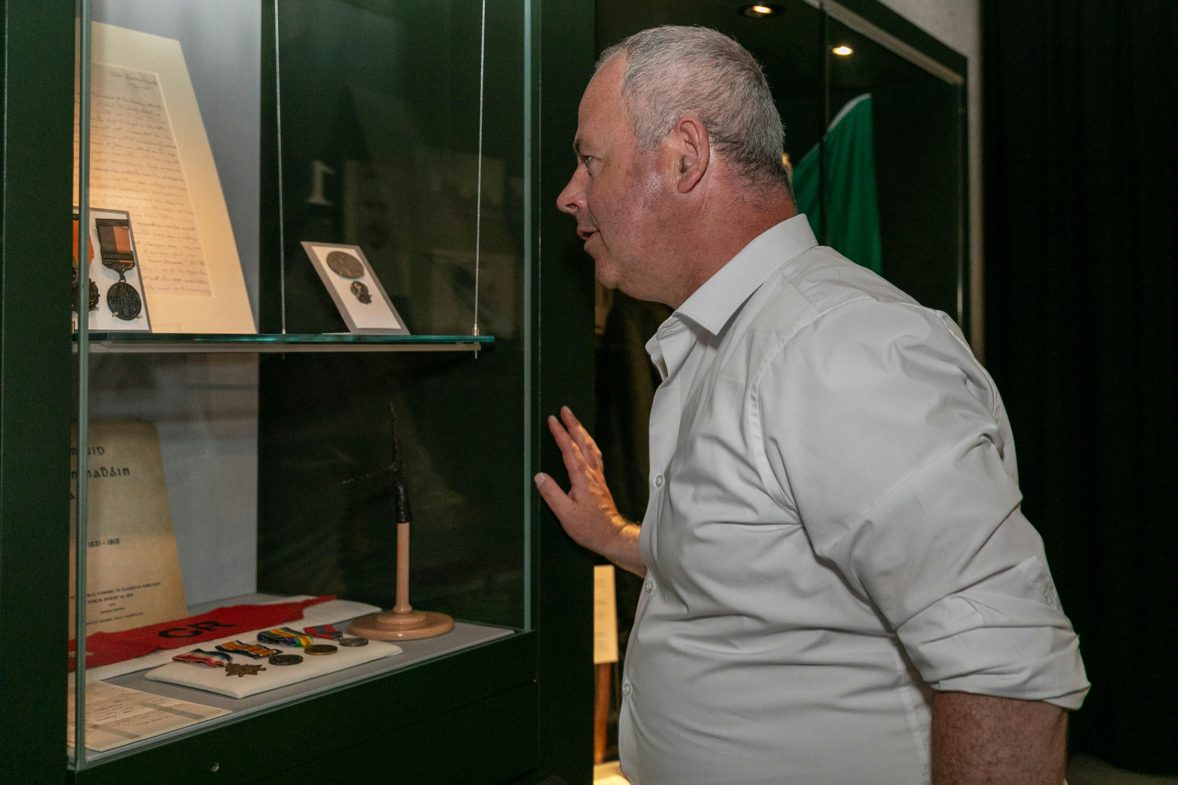 Ein Mann in einem weißen Hemd betrachtet Ausstellungsstücke in einem Museum, darunter Medaillen und gerahmte Fotos.