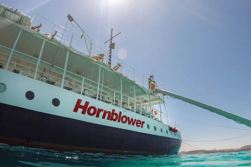 A close-up of the side of a ship named 'Hornblower' floating on clear turquoise water under a sunny sky.