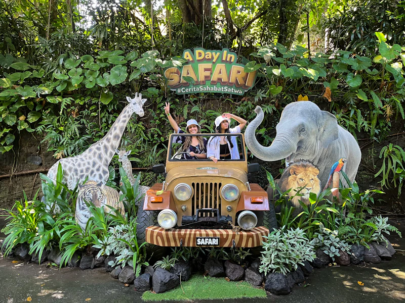 Two people pose in a safari-themed setup with a jeep surrounded by cutouts of animals and greenery, under a sign "A Day in Safari."