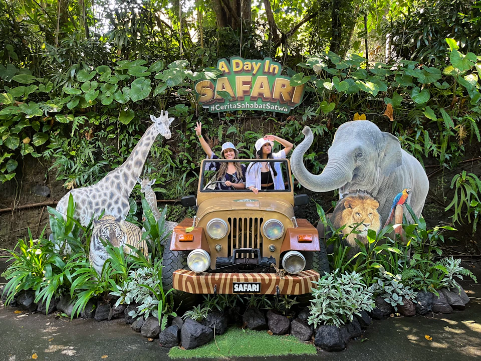 Two people pose in a safari-themed setup with a jeep surrounded by cutouts of animals and greenery, under a sign "A Day in Safari."