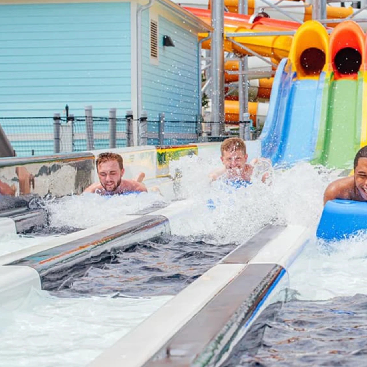 People racing down water slides on mats in a water park, splashing into the pool at the bottom with colorful slides in view.