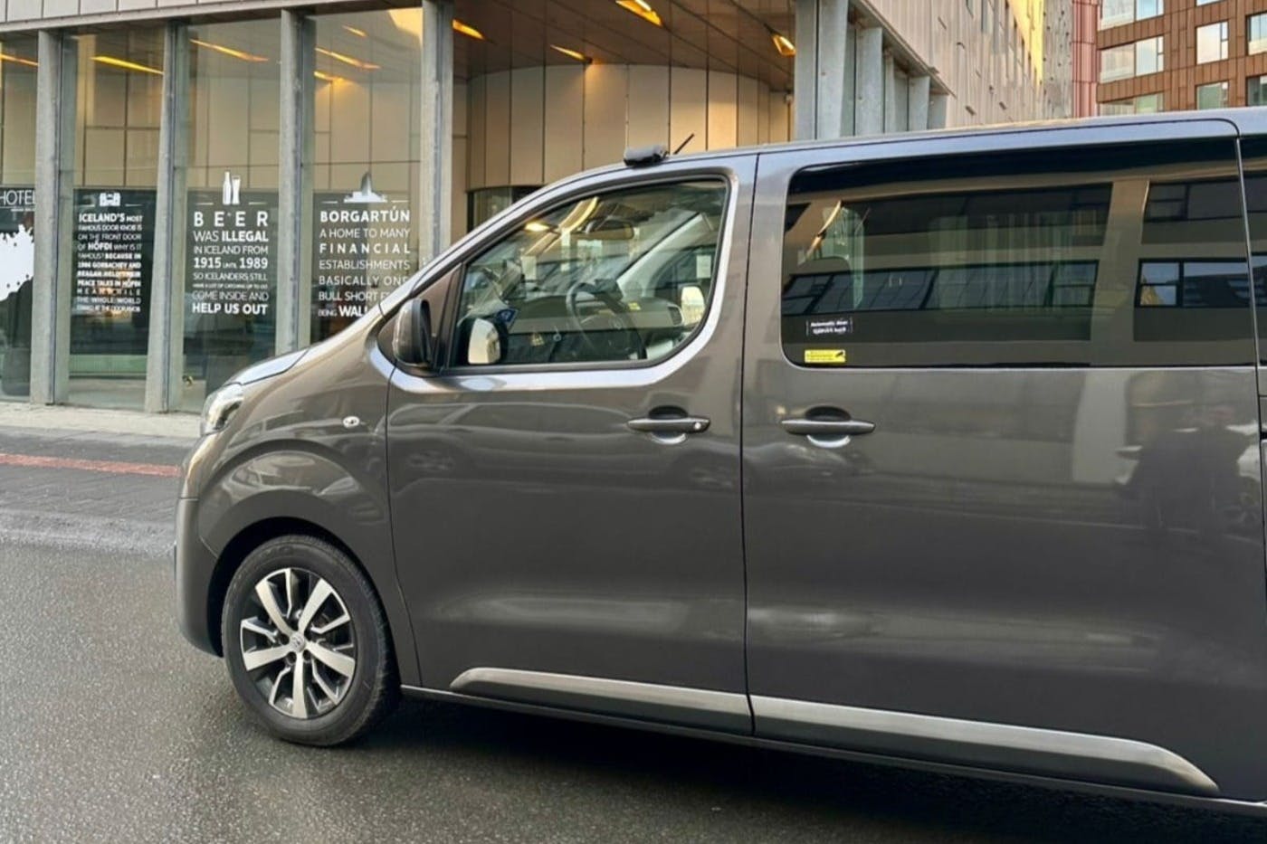 Side view of a gray van parked on a street, with a building featuring large windows and visible text in the background.