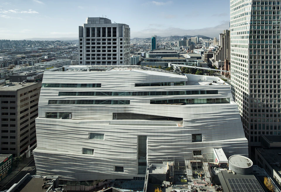 Modern white building with wavy façade set amidst cityscape with surrounding high-rises and distant mountains under a cloudy sky.