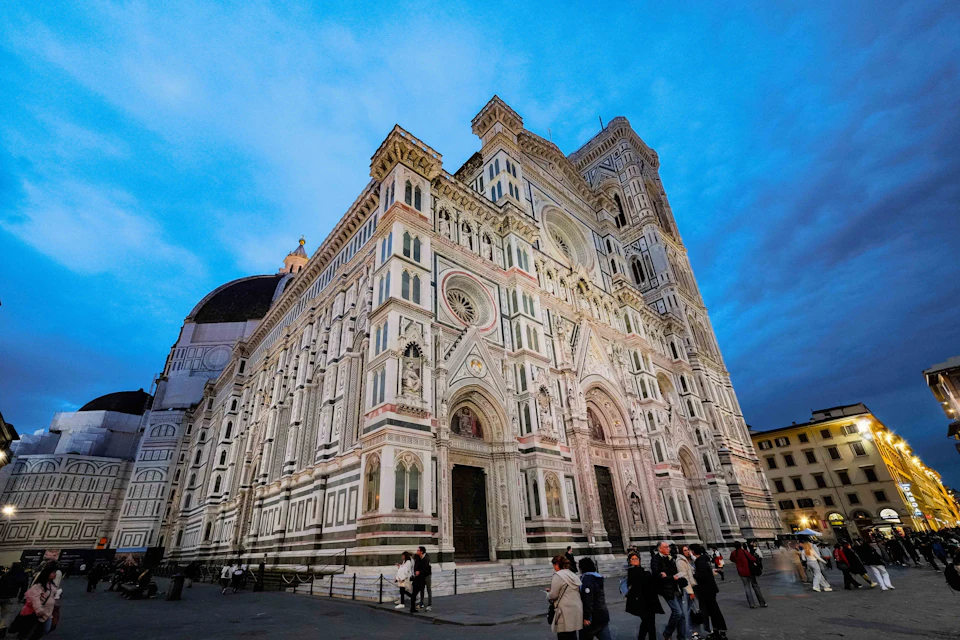 People walk near a large, ornate cathedral with intricate details and a tall dome under a twilight sky.
