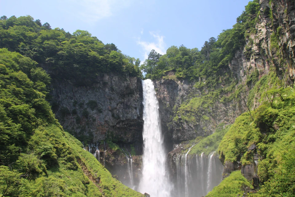 A tall waterfall cascading down a rocky cliff surrounded by lush green foliage under a clear blue sky.