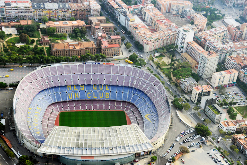 Aerial view of a large stadium with "MÉS QUE UN CLUB" seats, surrounded by urban buildings and streets.