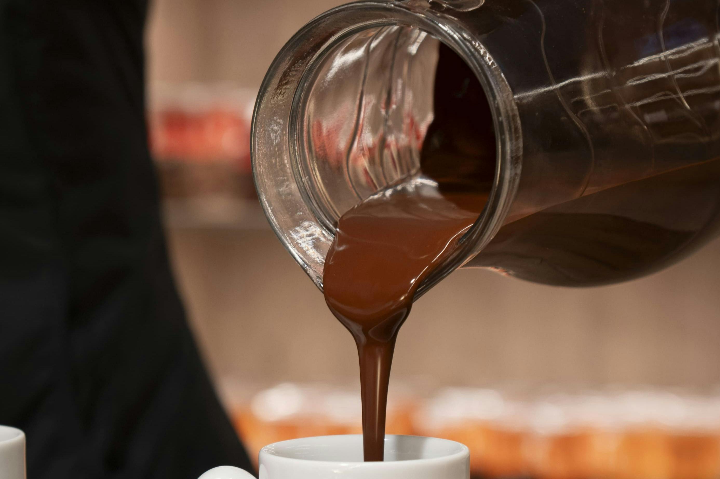 Gros plan d'un filet de chocolat chaud s'écoulant d'un pichet en verre dans une tasse blanche.