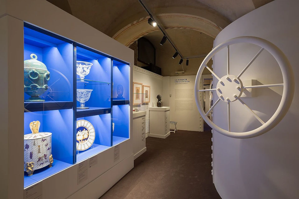 A museum exhibit featuring glassware in blue-lit cases, framed artwork on the walls, a sculpture, and a large wheel installation.