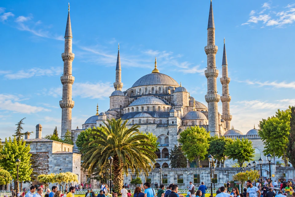 A grand mosque with multiple domes and minarets under a clear blue sky, surrounded by trees and people in a park.