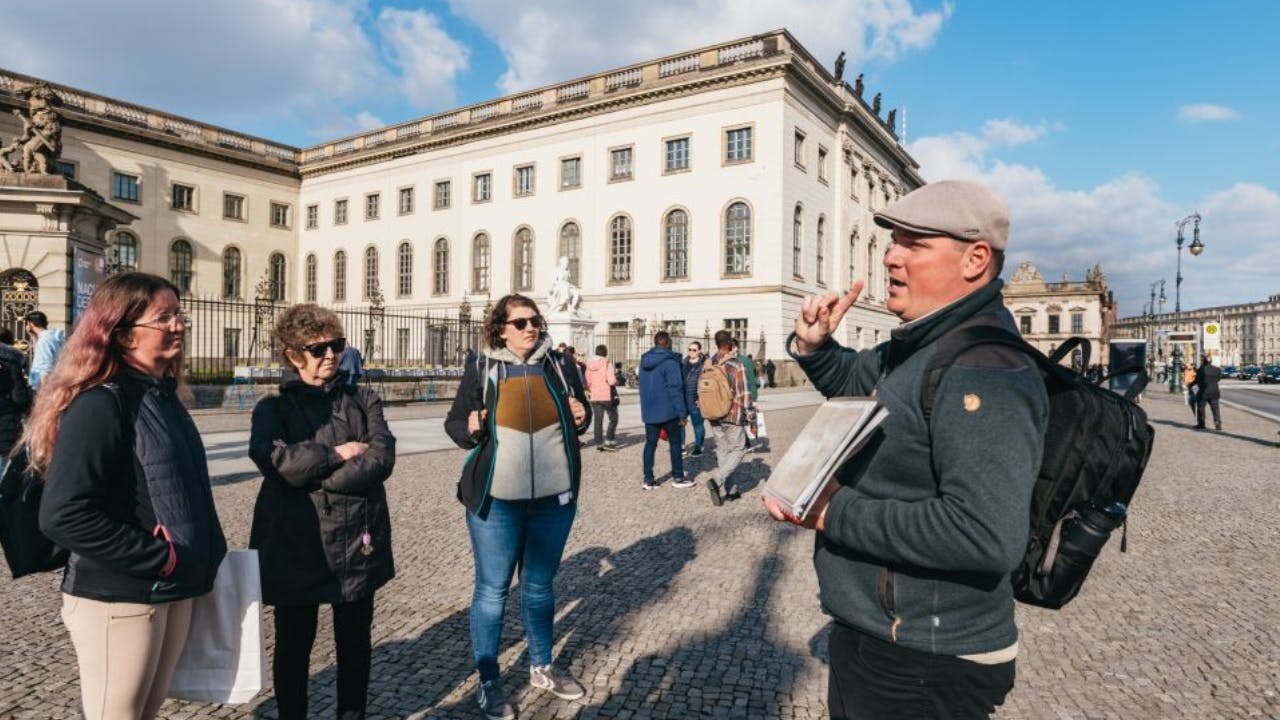 A man with a backpack and cap speaks to a group in front of an ornate building under a blue sky.