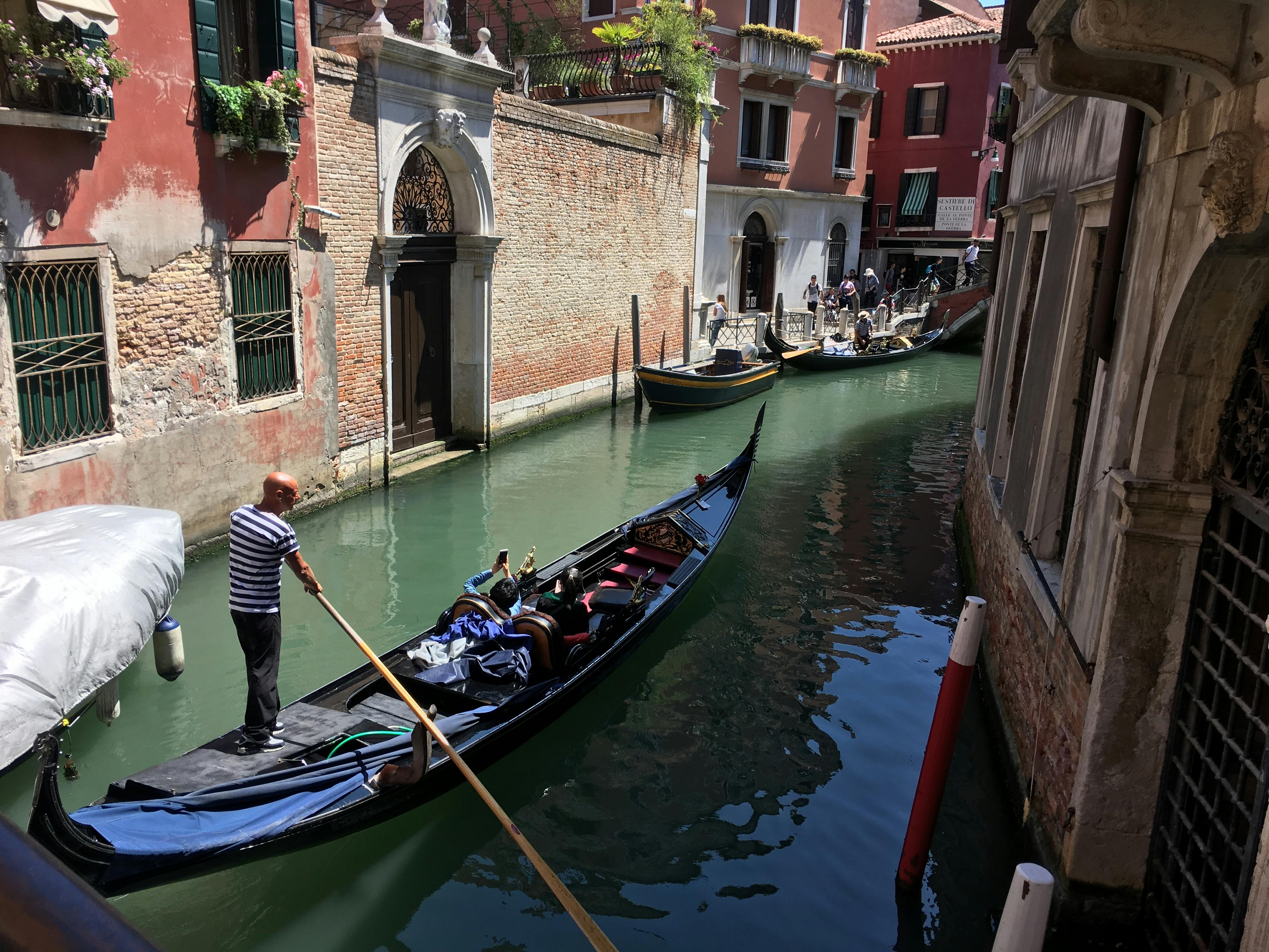 由船夫驾驶的贡多拉在意大利威尼斯的一条狭窄运河上航行，运河两旁环绕着乡村建筑。