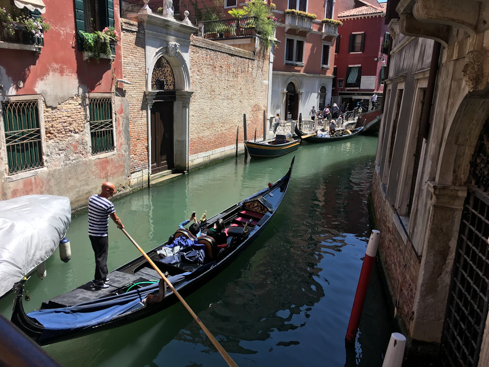 A gondolier navigates a gondola along a narrow canal lined with historic buildings in Venice, Italy, with other gondolas in the background.