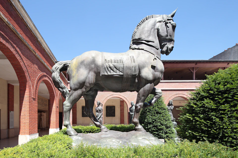 Large horse statue in a courtyard with arched brick colonnade, bushes, and a clear blue sky.