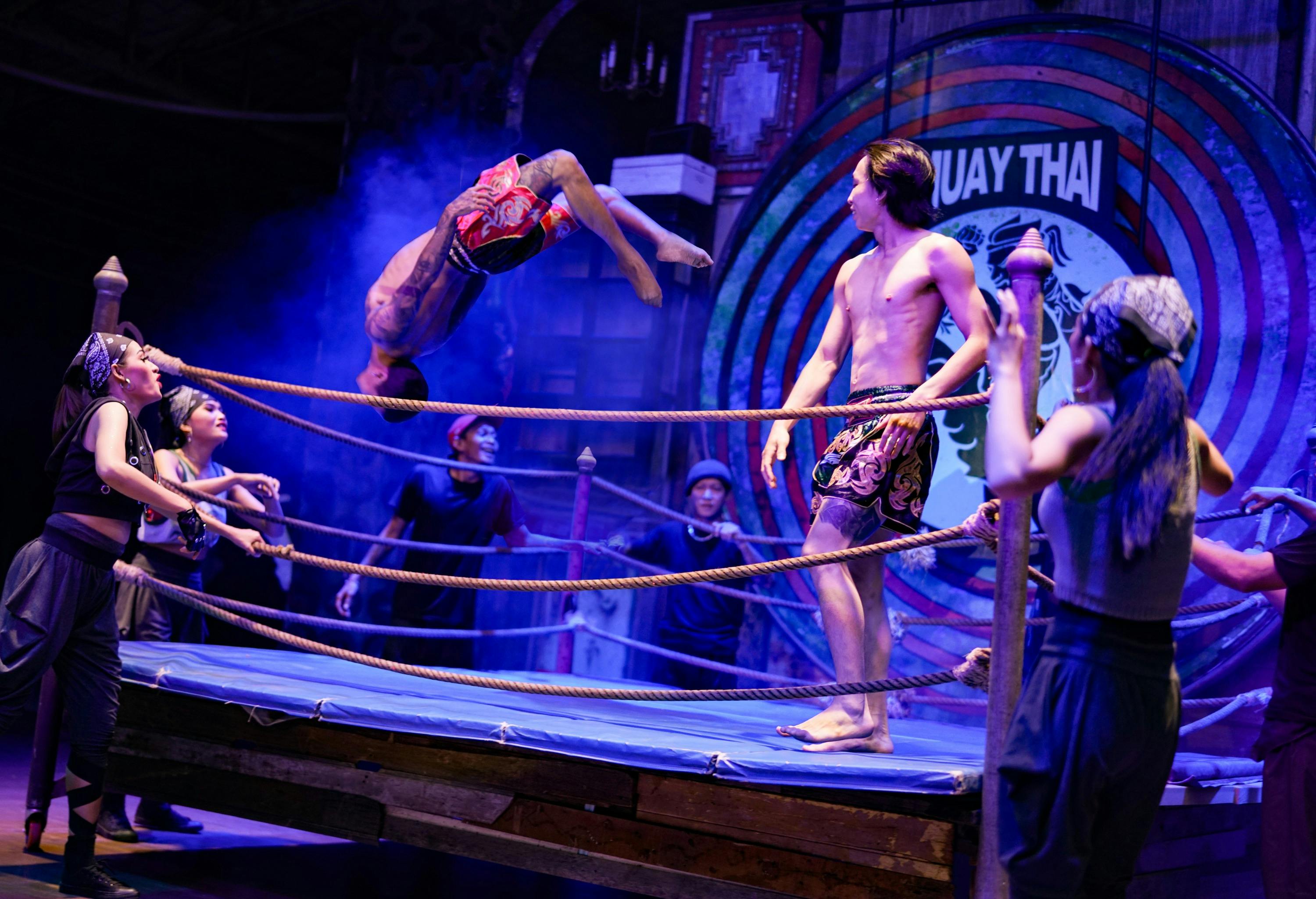 Występ Muay Thai na ringu z bokserem wykonującym salto w tył i innymi oglądającymi pod dramatycznym oświetleniem.