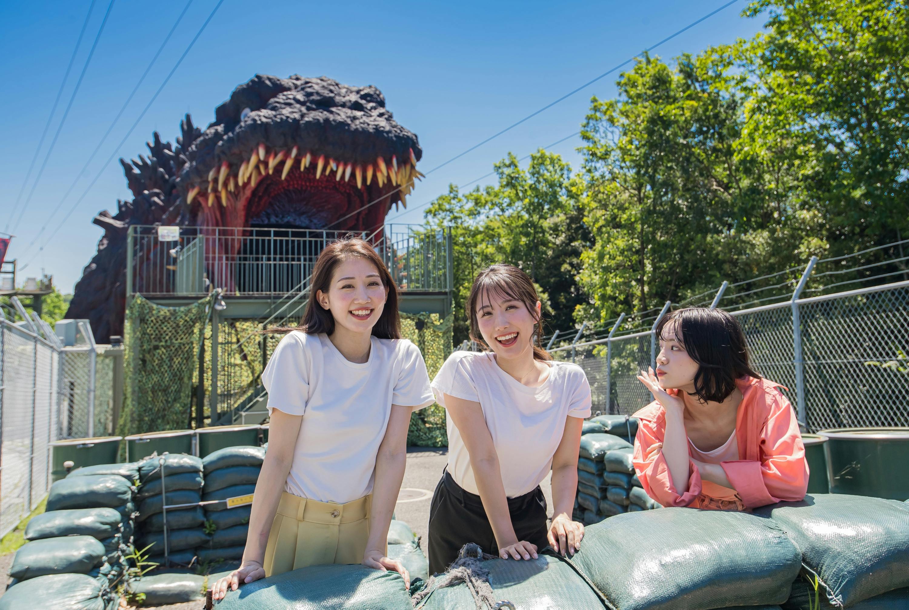 Three women smiling and posing near sandbags with a large dinosaur head structure in the background.