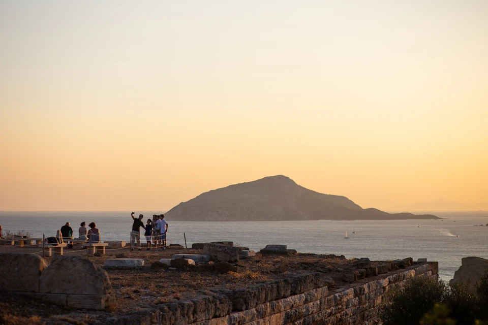 Sunset at Cape Sounio with people admiring the view.
Visitors enjoy golden hour as the sun sets behind a mountain and sea.